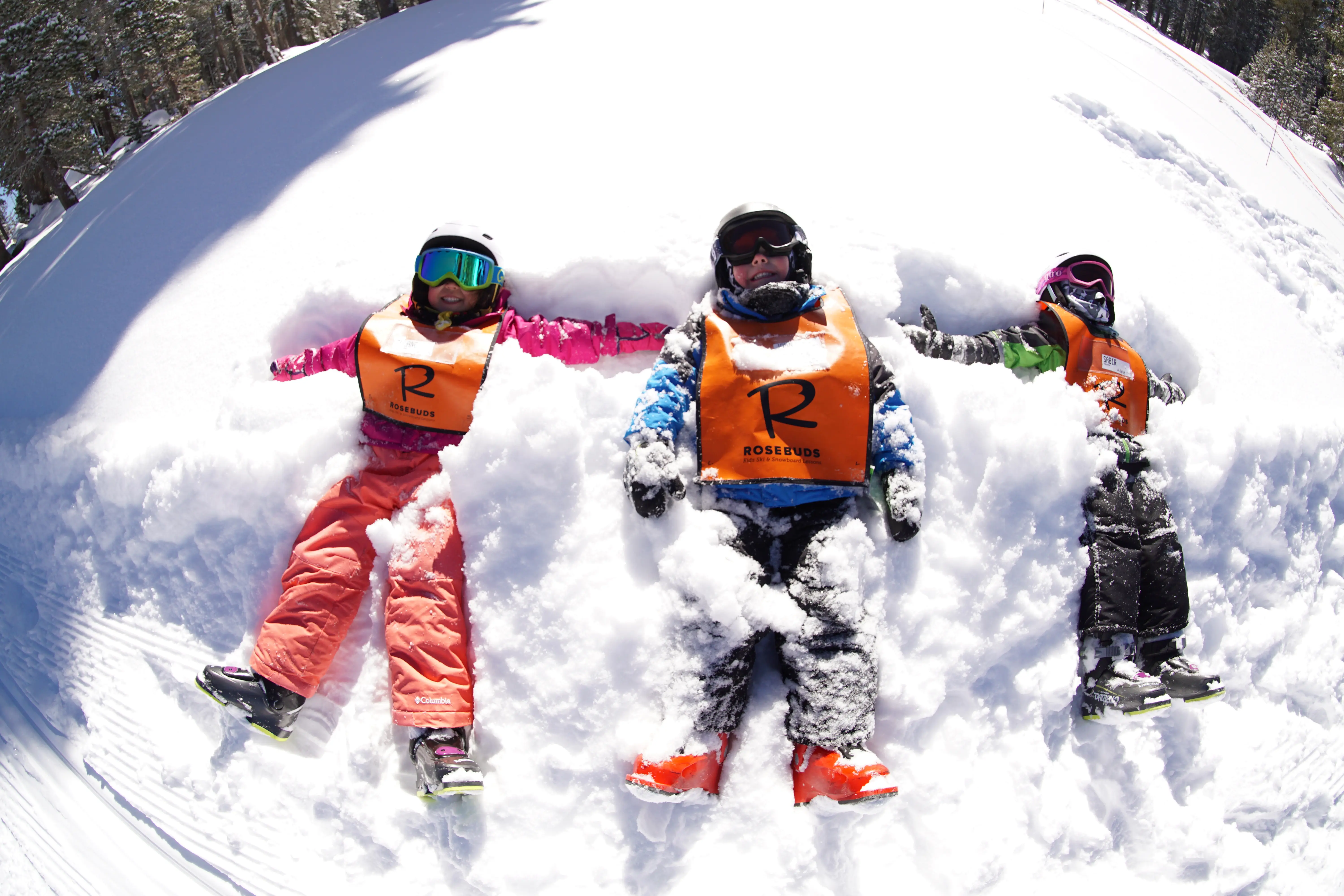 Three children wearing orange safety vests and snow gear lie on their backs in the snow, making snow angels, smiling and holding hands, with bright sunlight and a snowy landscape around them. Mt. Rose Ski Tahoe