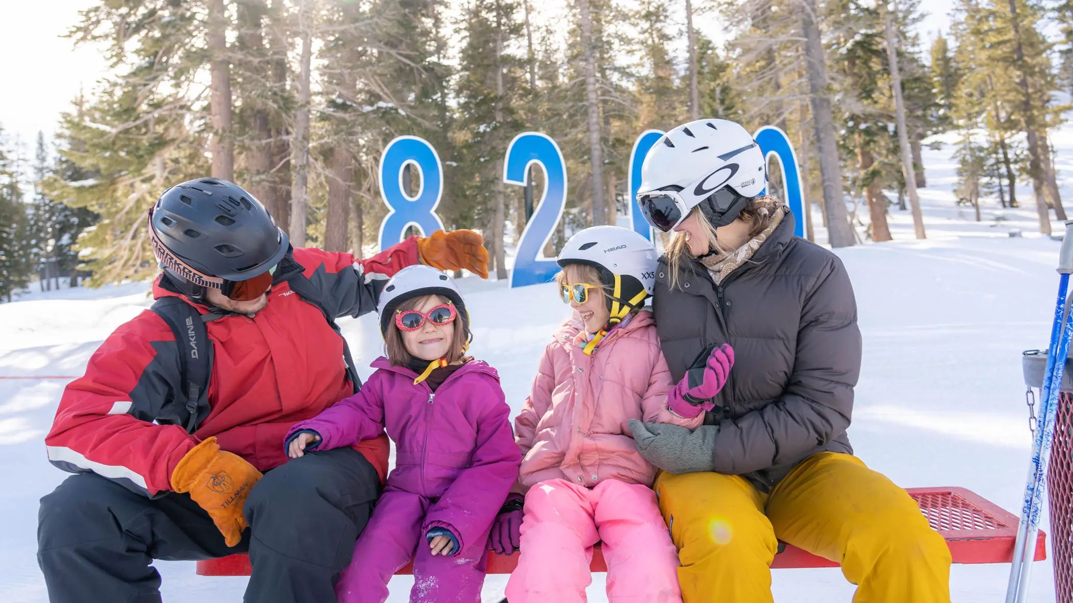 A group of four people, including children and adults, are skiing down a snowy slope on a sunny day. They are all wearing winter gear, including helmets, jackets, and ski boots. There are colorful markers on the snow from the best kids ski lessons, tall pine trees, and a clear blue sky in the background. Mt. Rose Ski Tahoe