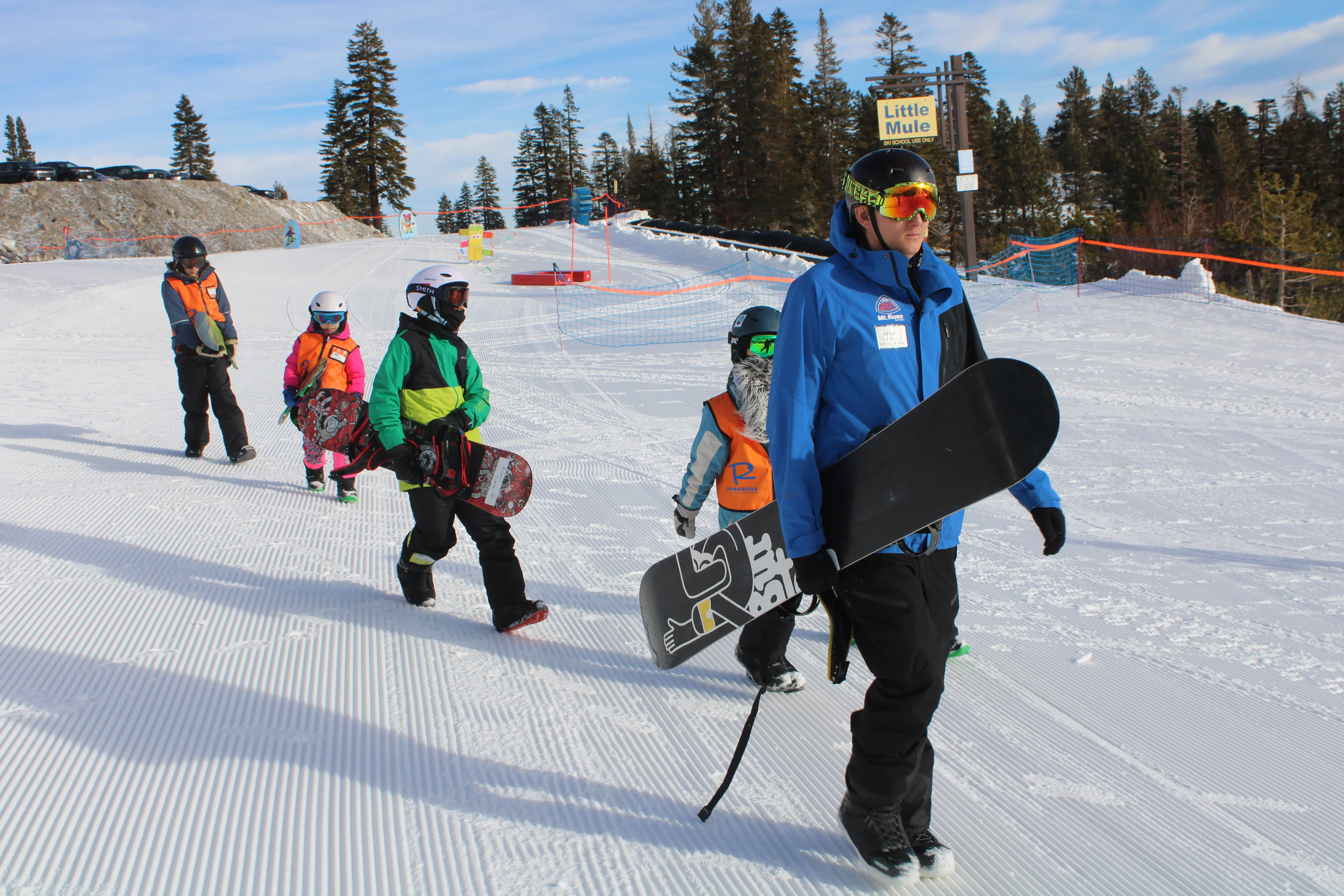 A snowboard instructor leads a group of children carrying snowboards on a snowy slope, with trees and a "Little Mule" sign in the background. The children wear helmets, goggles, and colorful winter gear. Mt. Rose Ski Tahoe