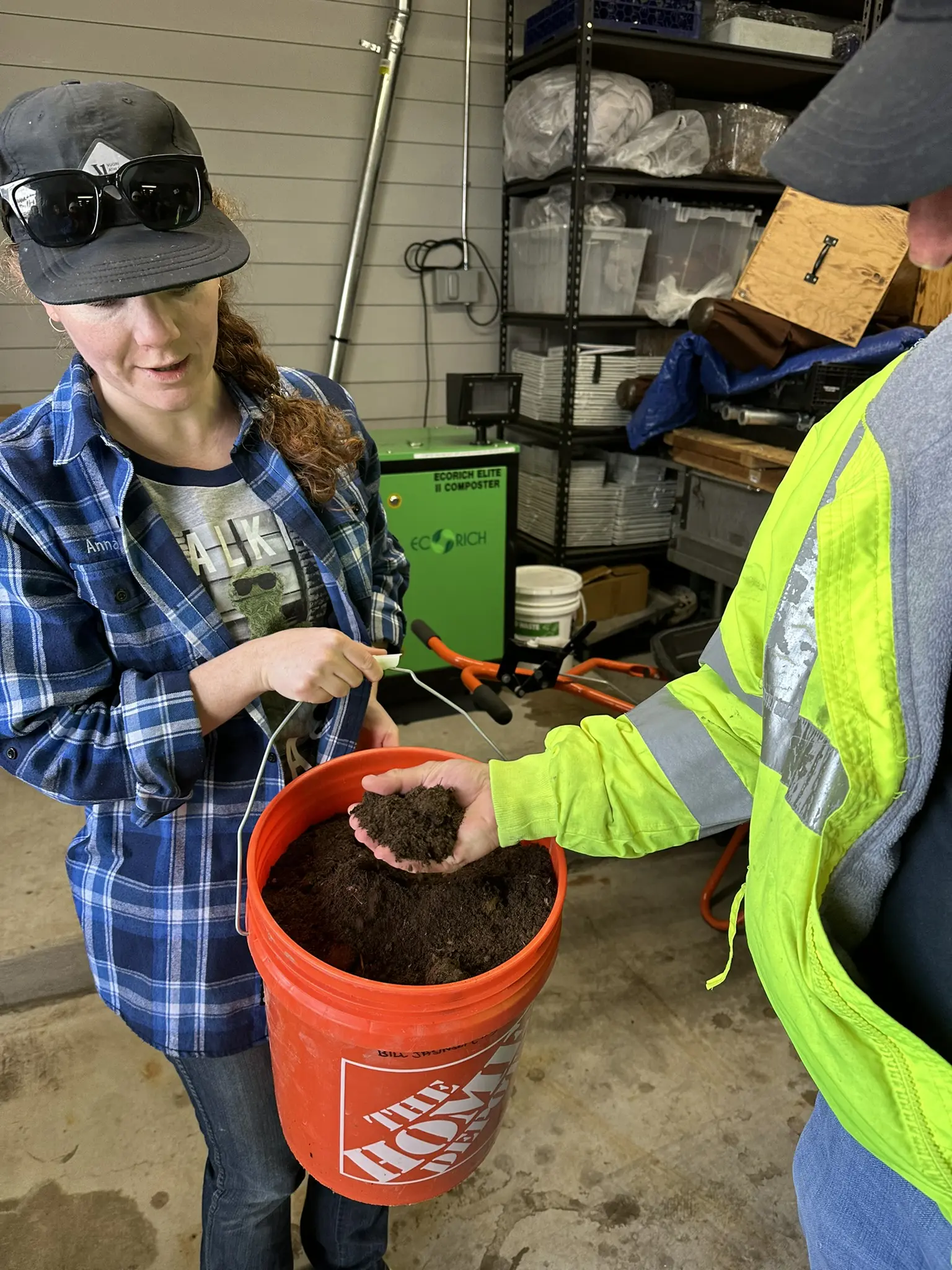 Two people stand in a garage holding a Home Depot bucket filled with dark soil; one person scoops soil with a trowel while the other holds out a handful. Various tools and supplies are visible in the background. Mt. Rose Ski Tahoe