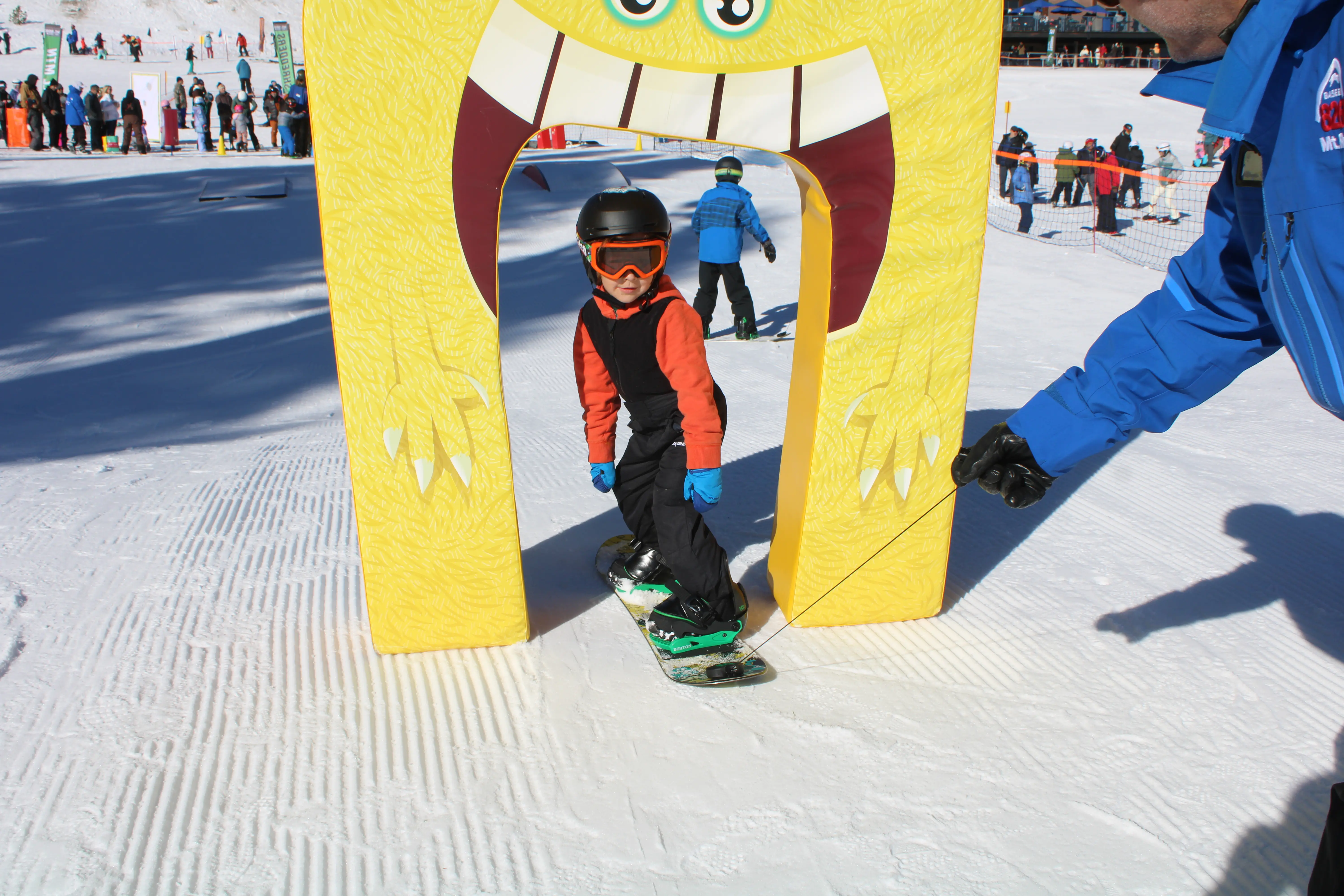 A child in snowboarding gear rides under a yellow arch shaped like a monster face on a snowy slope, being guided by an adult holding a strap. People and a ski area are visible in the background. Mt. Rose Ski Tahoe