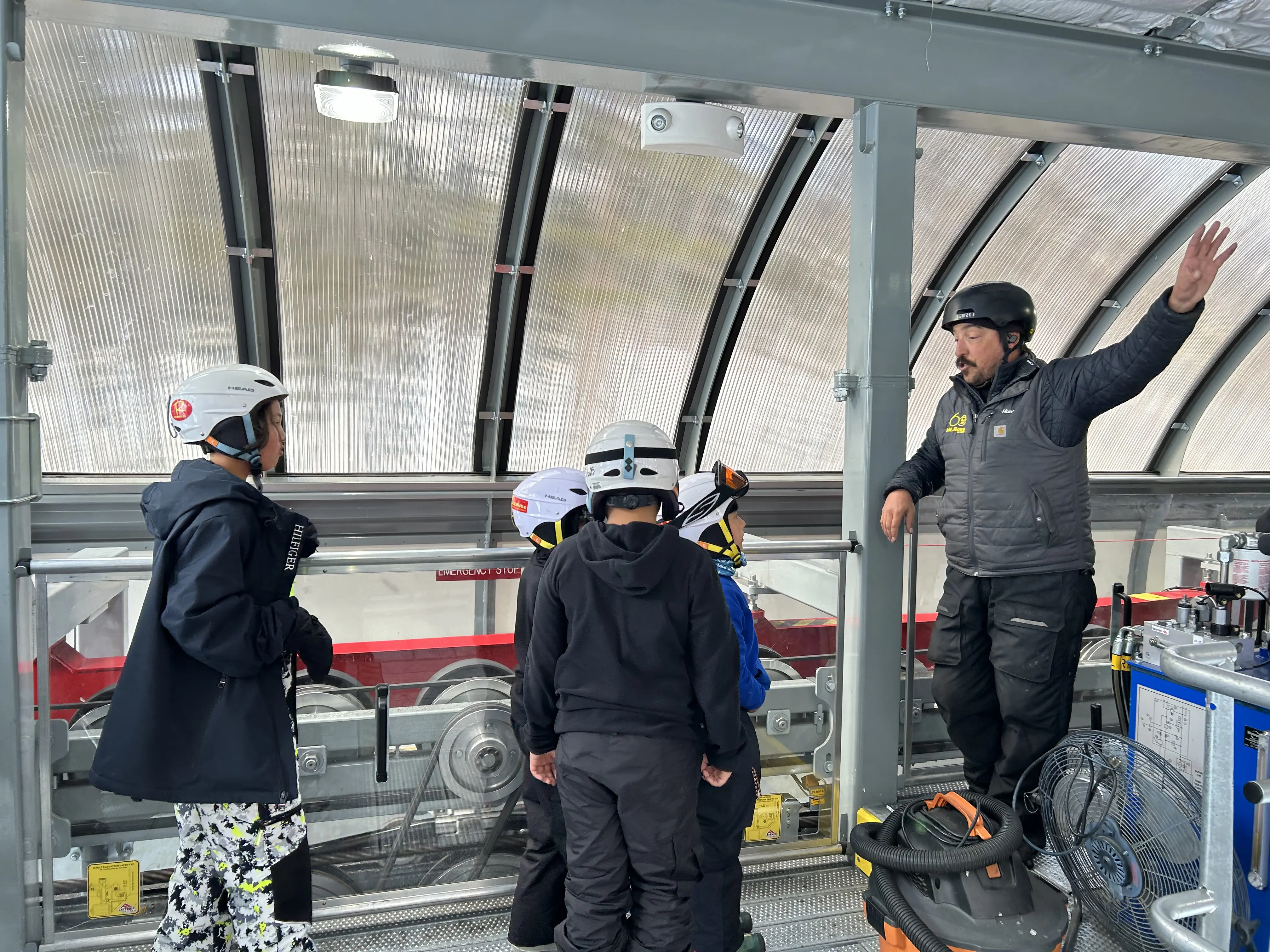 A group of five people wearing helmets and winter gear stand inside a ski lift terminal; one adult gestures while instructing the others, who appear to be listening attentively. Mt. Rose Ski Tahoe
