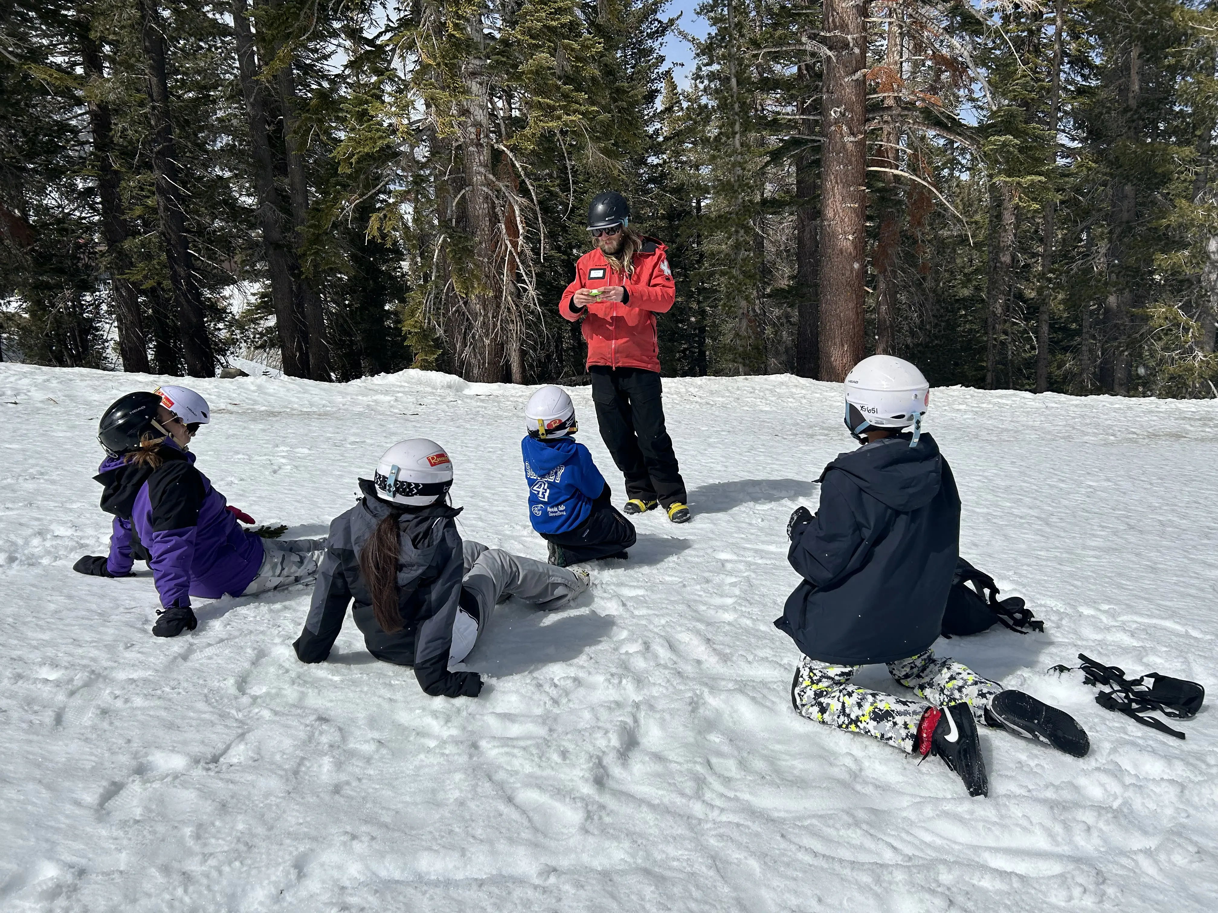 A ski instructor in a red jacket teaches four people sitting on snow, all wearing helmets and winter gear, with a backdrop of tall pine trees. Mt. Rose Ski Tahoe
