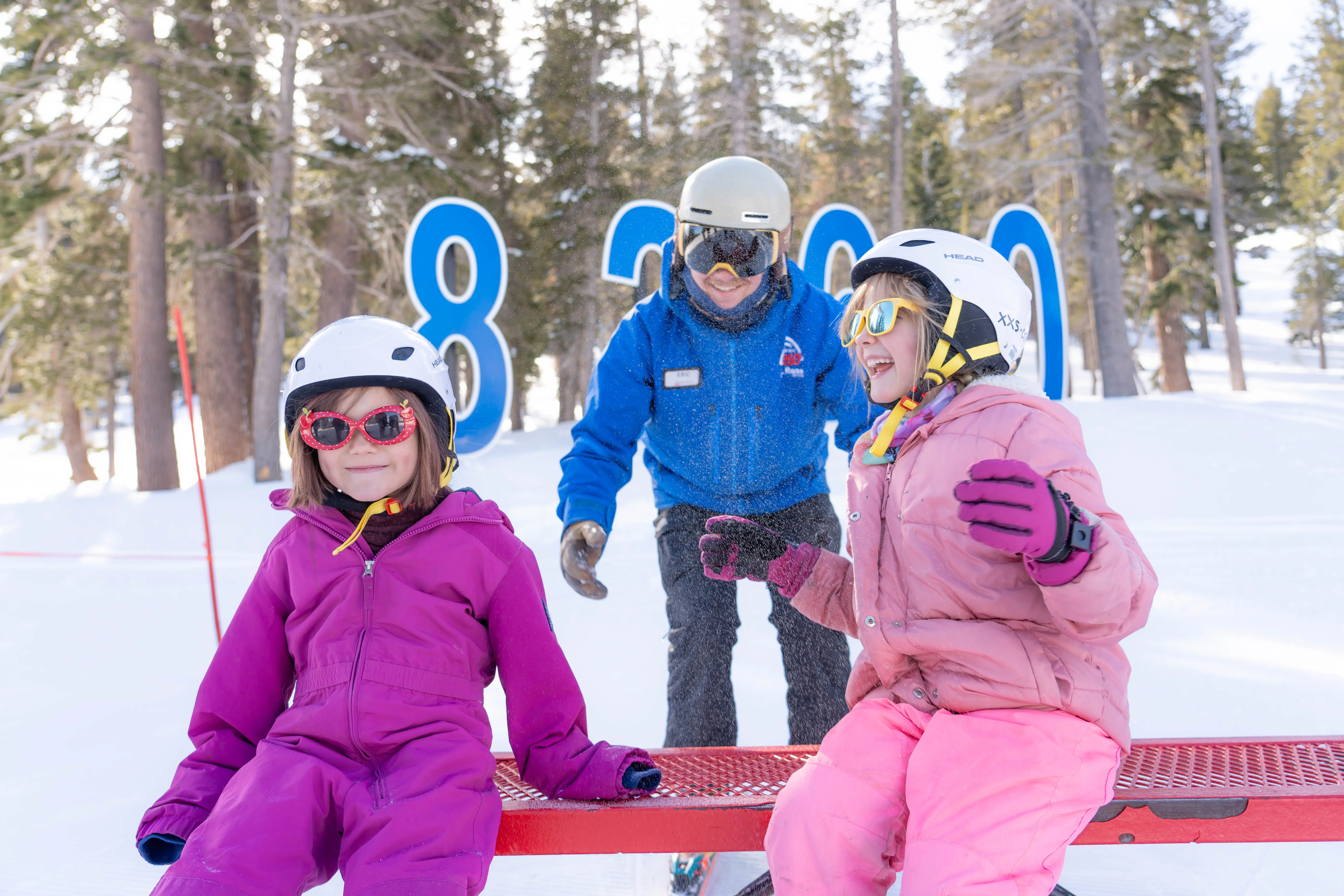 Two young children in colorful snowsuits and helmets sit on a red bench in the snow, smiling and laughing, while an adult in blue ski gear stands behind them. Snow-covered trees and large blue numbers are in the background. Mt. Rose Ski Tahoe