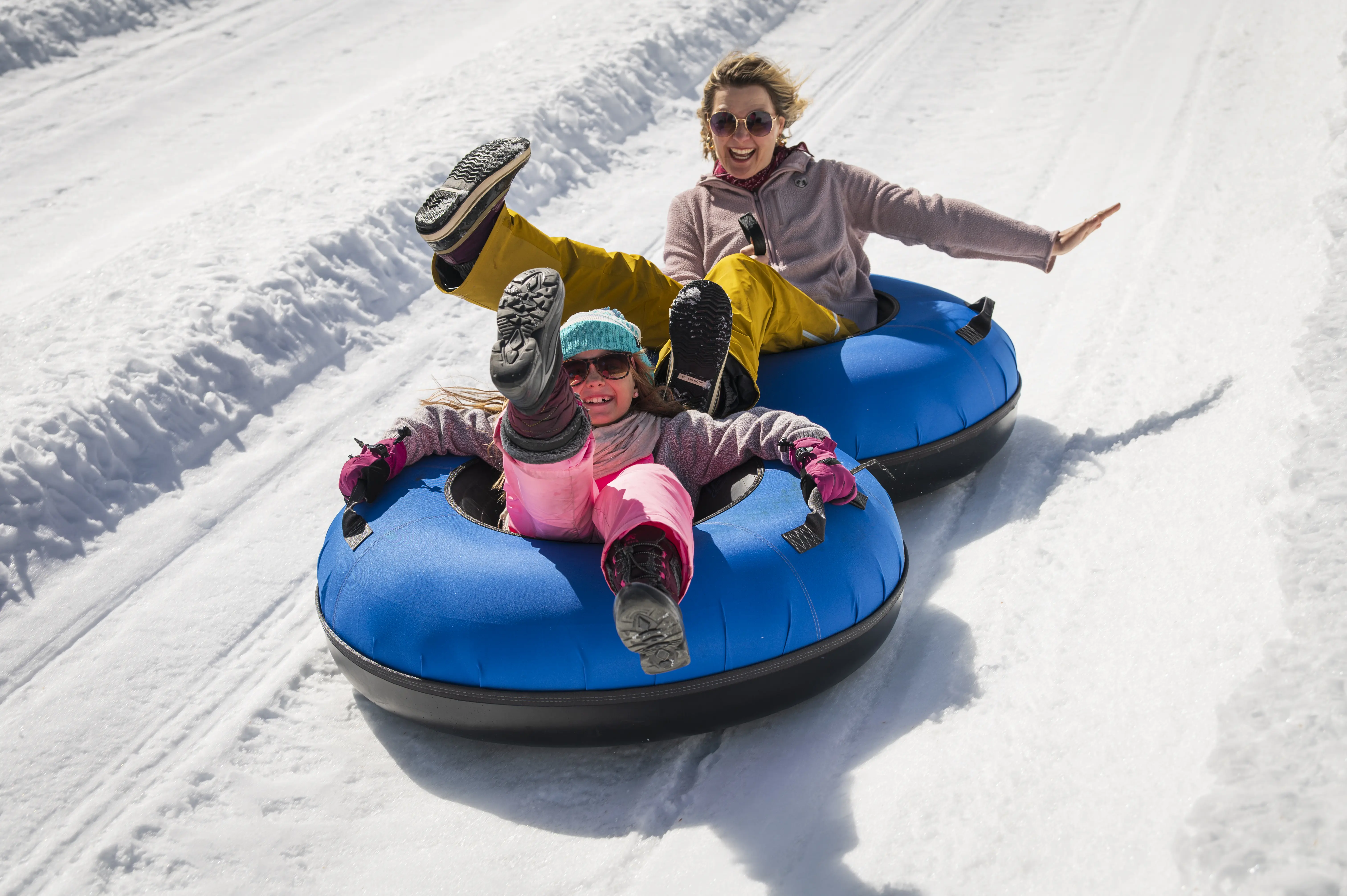 Two people, an adult and a child, laugh and raise their legs while riding blue snow tubes down a snowy hill on a sunny day. Both are dressed in winter clothes and appear to be enjoying the ride. Mt. Rose Ski Tahoe