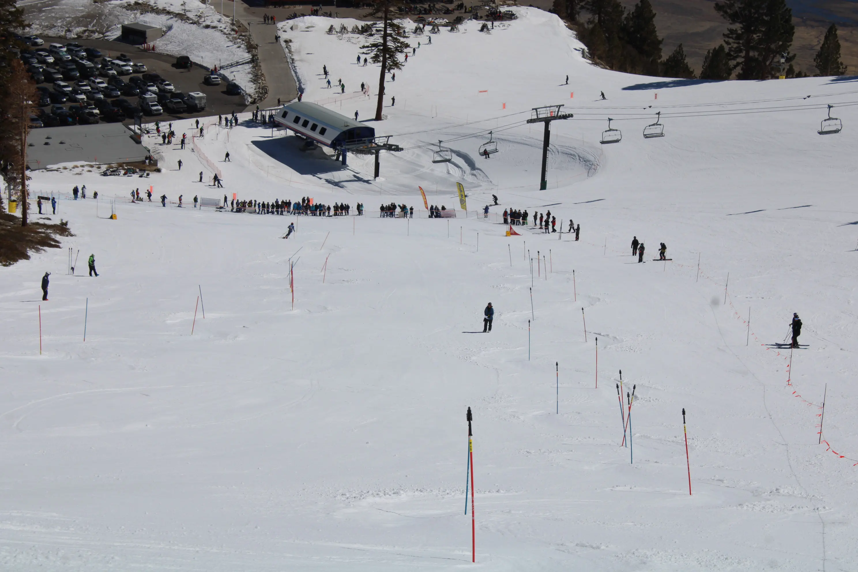 A snowy ski slope with skiers descending, lift chairs overhead, and people waiting in line near a ski lift station. Trees and parked cars are visible in the background. Mt. Rose Ski Tahoe