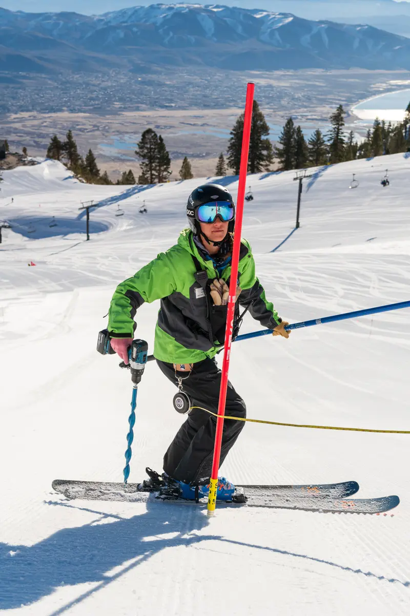 A person in a green and black jacket skis on a snowy slope, holding a pole and an auger drill. The background shows mountains, trees, and a valley under a clear sky. Mt. Rose Ski Tahoe