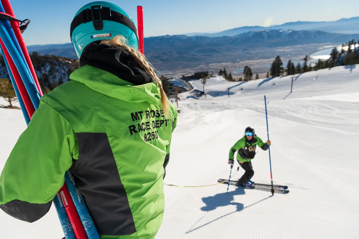 Two people on a snowy mountain slope, one standing with ski poles and wearing a jacket labeled "MT ROSE RACE DEPT," and another skiing downhill, with mountains and a lake in the background. Mt. Rose Ski Tahoe