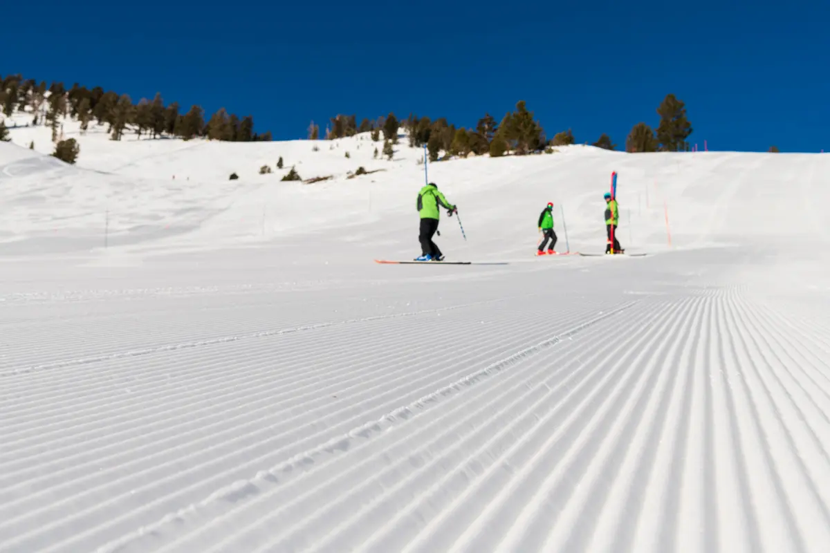 Three skiers in green jackets are on a freshly groomed snowy ski slope under a clear blue sky, with trees and snowy hills in the background. Mt. Rose Ski Tahoe
