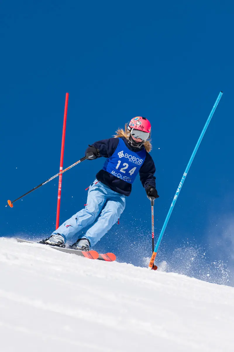 A skier in a pink helmet and blue bib number 124 navigates between slalom gates on a snowy slope, kicking up snow under a clear blue sky. Mt. Rose Ski Tahoe