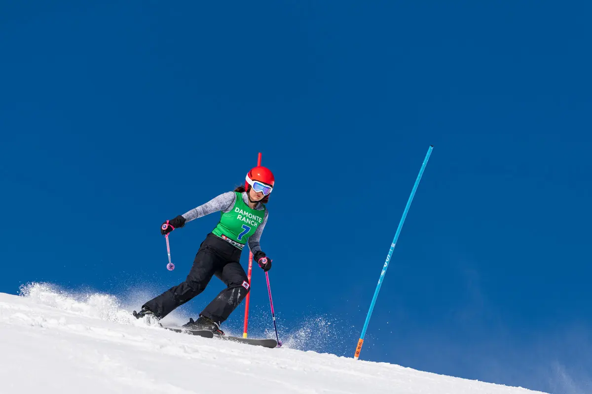 A skier wearing a red helmet, green bib, and goggles races downhill, navigating between brightly colored slalom poles on a sunny, clear day with a deep blue sky. Mt. Rose Ski Tahoe