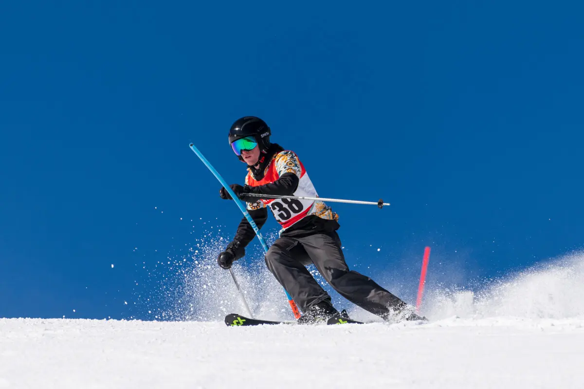 A skier wearing a helmet, goggles, and a numbered vest navigates a slalom course on a snowy slope, kicking up snow as they turn, with a clear blue sky in the background. Mt. Rose Ski Tahoe
