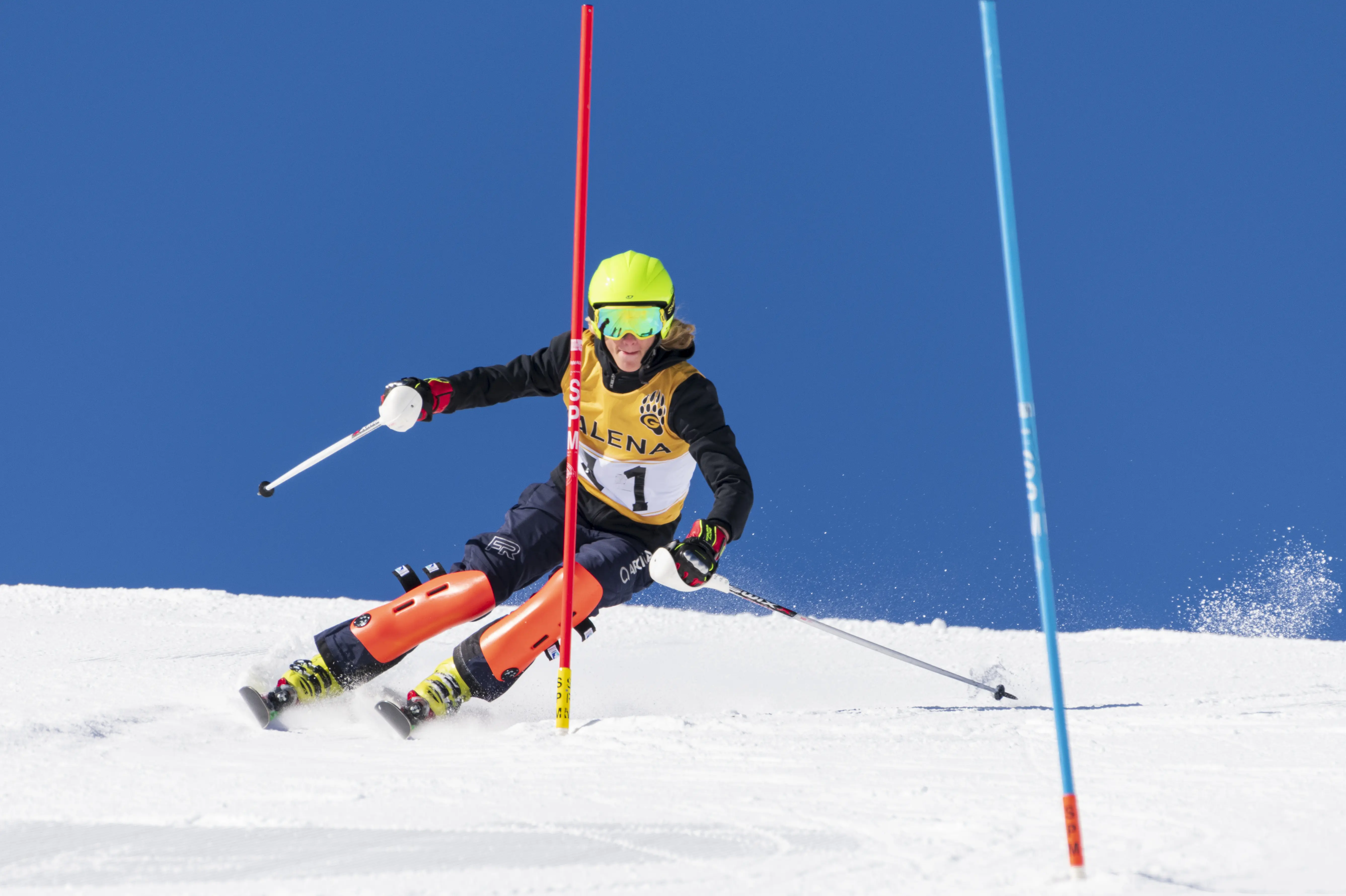 A skier in a yellow bib and green helmet races downhill in a High School Racing event, navigating between slalom poles on a snowy slope under a clear blue sky. Mt. Rose Ski Tahoe