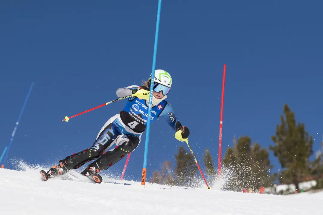 A skier in a helmet and goggles races downhill through slalom gates on a snowy slope, kicking up snow with trees and a clear blue sky in the background. Mt. Rose Ski Tahoe