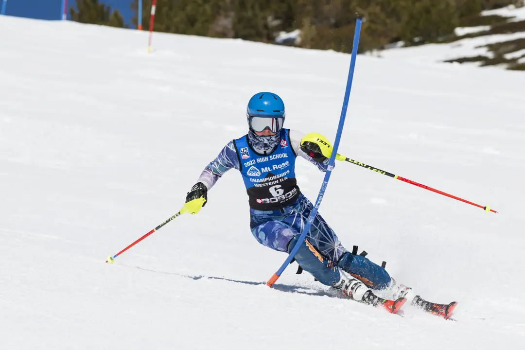 A skier in colorful gear and a blue helmet races downhill, leaning into a turn and navigating between slalom poles on a snowy slope during a ski competition. Mt. Rose Ski Tahoe