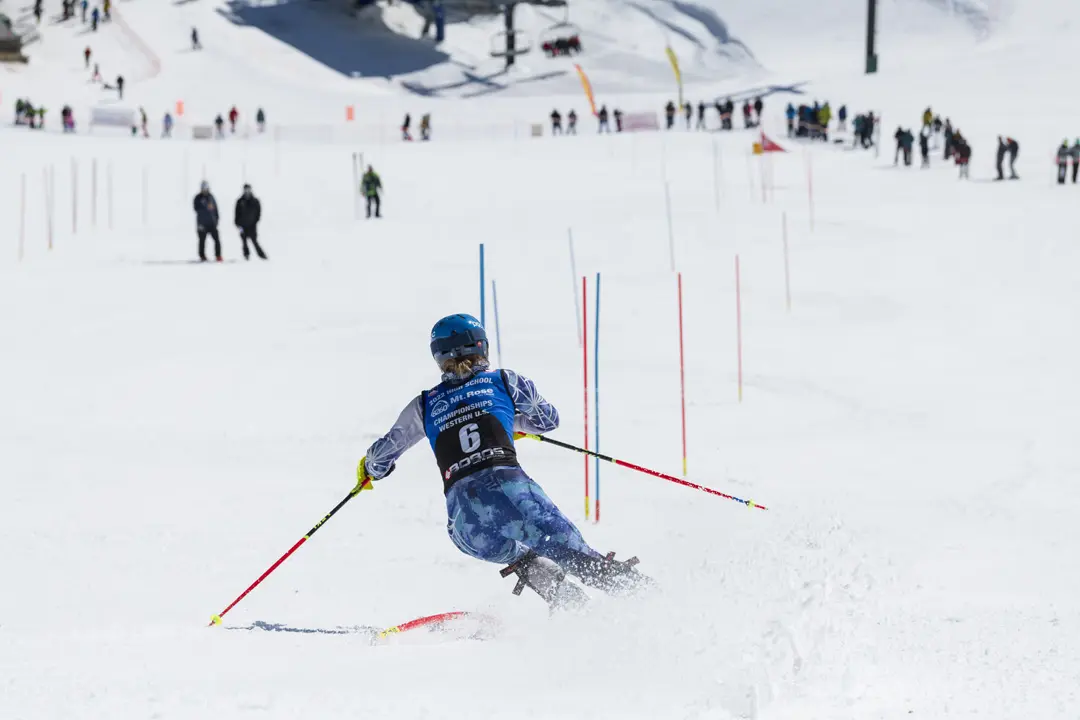 A skier wearing a blue helmet and competition bib number 6 navigates between red and blue slalom poles on a snowy slope, with spectators and other skiers visible in the background. Mt. Rose Ski Tahoe