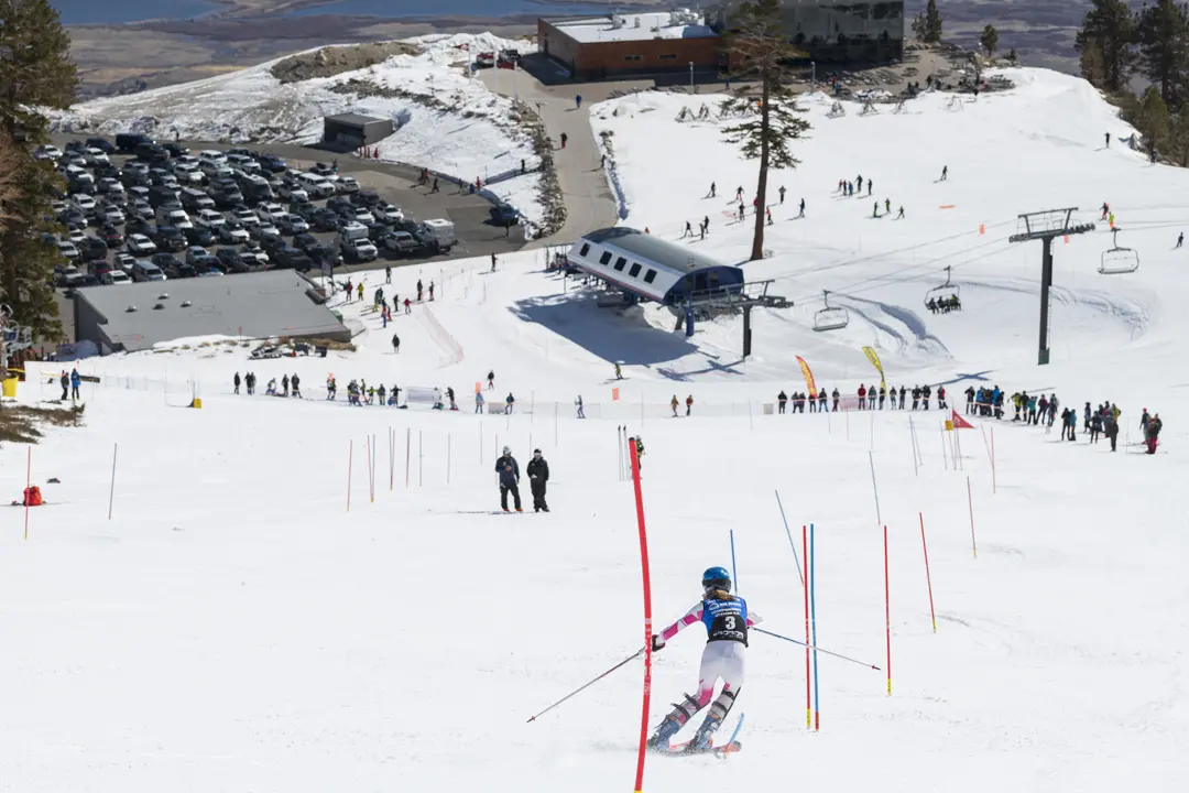 A skier in a race suit navigates a slalom course on a snowy mountain slope, with spectators, a ski lift, parked cars, and a lodge visible in the background under a clear sky. Mt. Rose Ski Tahoe
