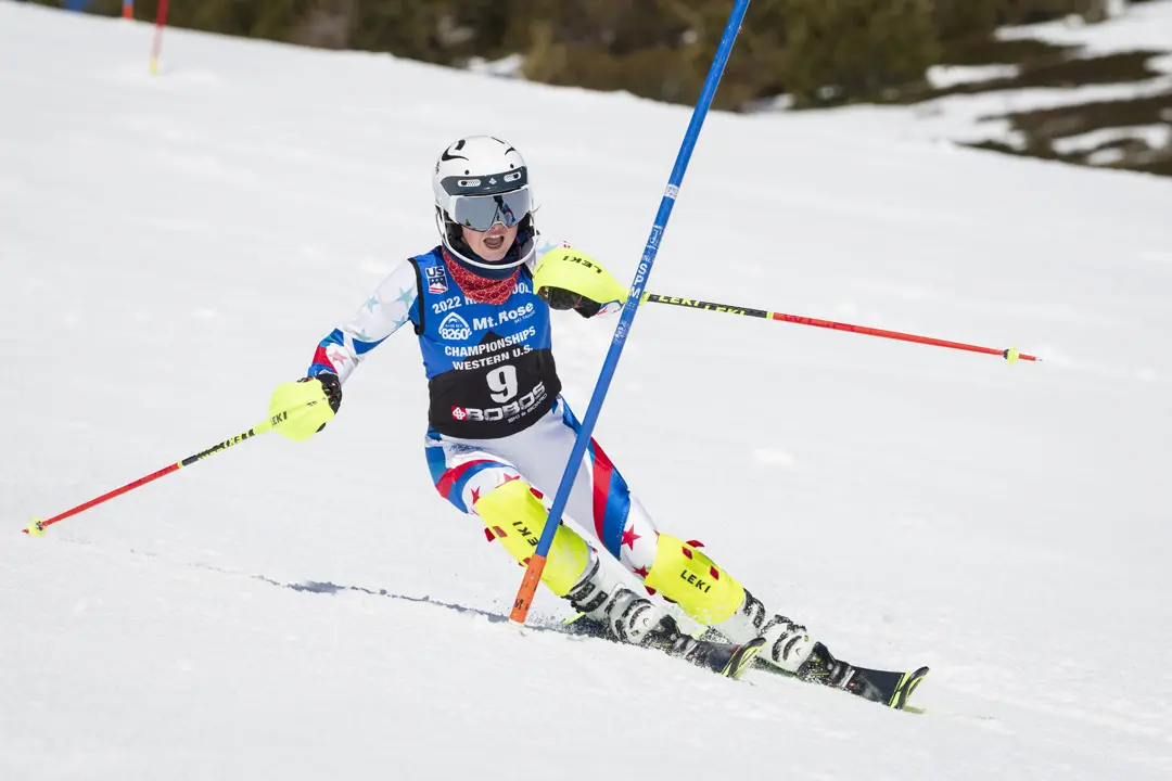 A skier wearing a helmet, goggles, and race suit is navigating a slalom course, leaning sharply while passing a gate on a snowy slope with some patches of brown in the background. Mt. Rose Ski Tahoe