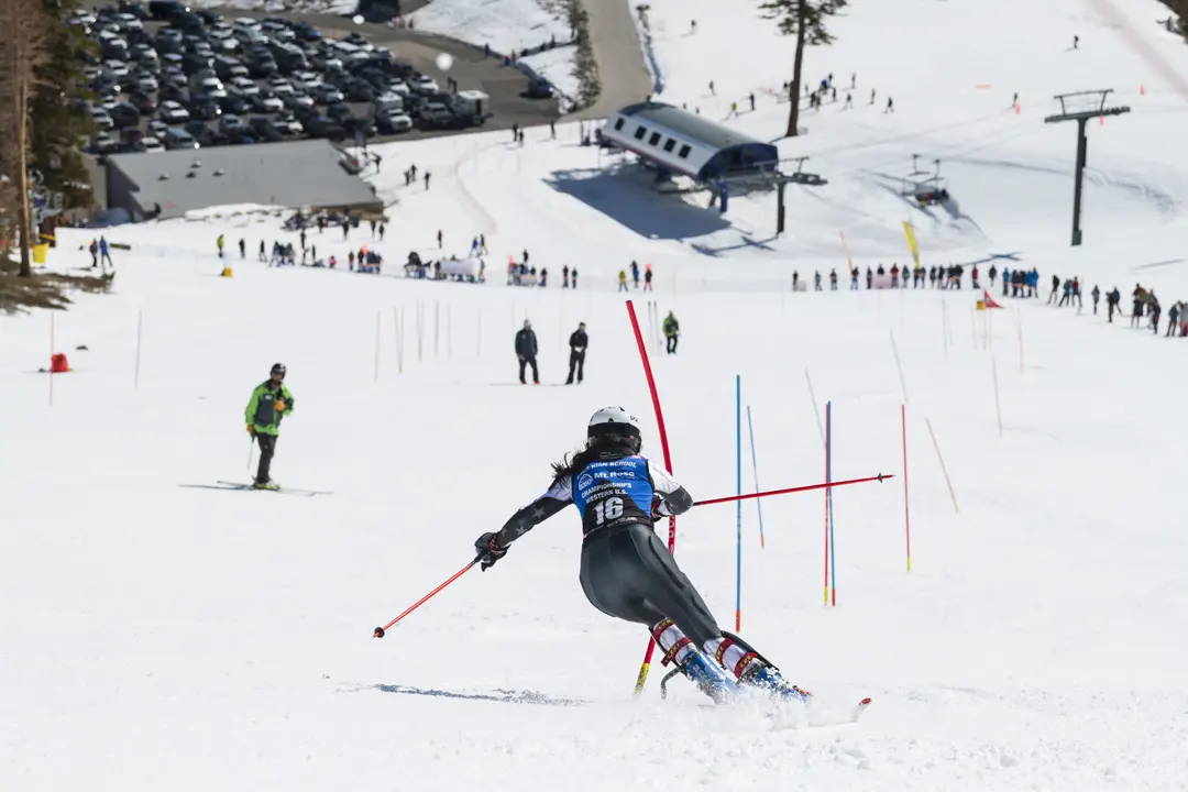 A skier wearing a helmet and race bib navigates through slalom poles on a snowy slope, with spectators, a ski lift, and cars visible in the background under a clear sky. Mt. Rose Ski Tahoe