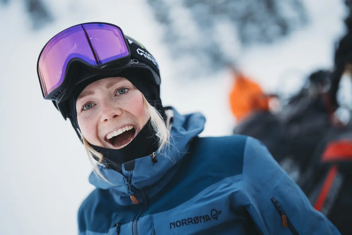 A smiling person in winter clothing and ski goggles stands outdoors in a snowy landscape, with blurred people and equipment in the background. Mt. Rose Ski Tahoe A smiling person in winter clothing and ski goggles stands outdoors in a snowy landscape, with blurred people and equipment in the background. Mt. Rose Ski Tahoe