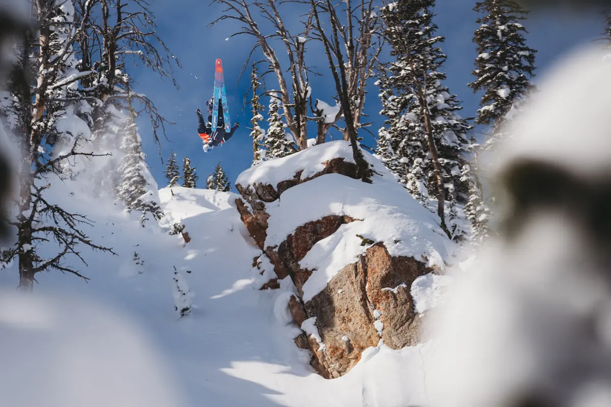 A skier performs a backflip off a snow-covered cliff surrounded by pine trees under a clear blue sky, with fresh powder and rocky outcrops visible in a winter mountain landscape. Mt. Rose Ski Tahoe A skier performs a backflip off a snow-covered cliff surrounded by pine trees under a clear blue sky, with fresh powder and rocky outcrops visible in a winter mountain landscape. Mt. Rose Ski Tahoe