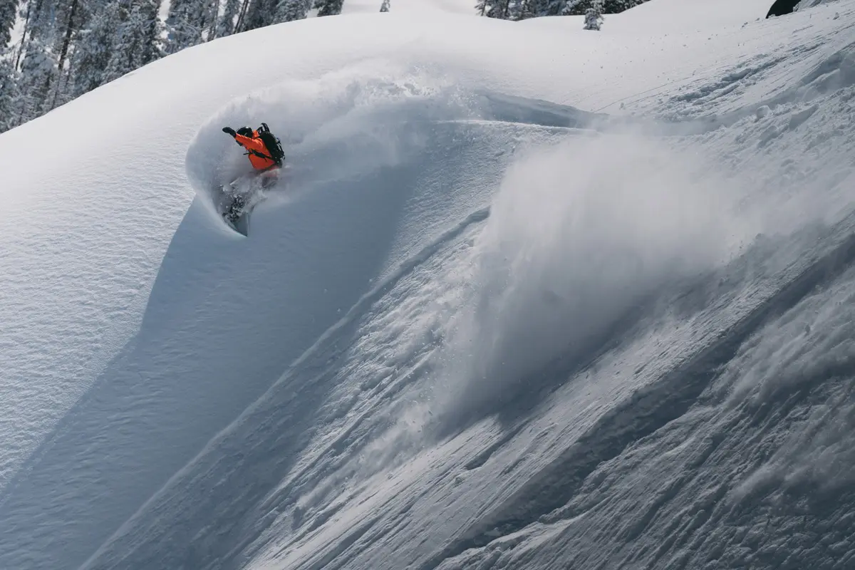 A snowboarder in an orange jacket carves through deep powder on a steep, snow-covered mountain slope, creating a dramatic spray of snow behind them under a clear sky. Mt. Rose Ski Tahoe A snowboarder in an orange jacket carves through deep powder on a steep, snow-covered mountain slope, creating a dramatic spray of snow behind them under a clear sky. Mt. Rose Ski Tahoe
