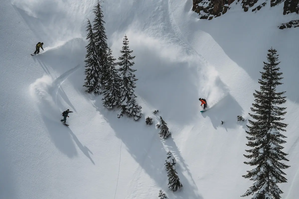 Three snowboarders carve down a snowy mountain slope, weaving between tall pine trees and leaving trails in fresh powder under bright daylight. Mt. Rose Ski Tahoe Three snowboarders carve down a snowy mountain slope, weaving between tall pine trees and leaving trails in fresh powder under bright daylight. Mt. Rose Ski Tahoe