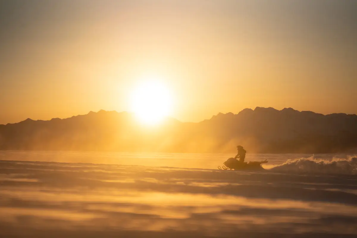 A person rides a snowmobile across a snowy landscape at sunset, with the sun low in the sky and mountains silhouetted in the background. Mt. Rose Ski Tahoe A person rides a snowmobile across a snowy landscape at sunset, with the sun low in the sky and mountains silhouetted in the background. Mt. Rose Ski Tahoe