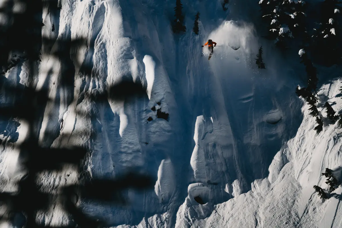 A skier in a bright orange jacket descends a steep, snow-covered mountain slope, surrounded by deep shadows and a few trees. Sunlight highlights the textured surface of the snow. Mt. Rose Ski Tahoe A skier in a bright orange jacket descends a steep, snow-covered mountain slope, surrounded by deep shadows and a few trees. Sunlight highlights the textured surface of the snow. Mt. Rose Ski Tahoe