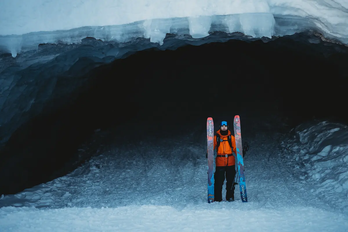 A person in an orange jacket stands on snowy ground in front of a large, dark ice cave, holding skis upright on either side. The cave has thick ice at the entrance, and snow covers the area around them. Mt. Rose Ski Tahoe A person in an orange jacket stands on snowy ground in front of a large, dark ice cave, holding skis upright on either side. The cave has thick ice at the entrance, and snow covers the area around them. Mt. Rose Ski Tahoe