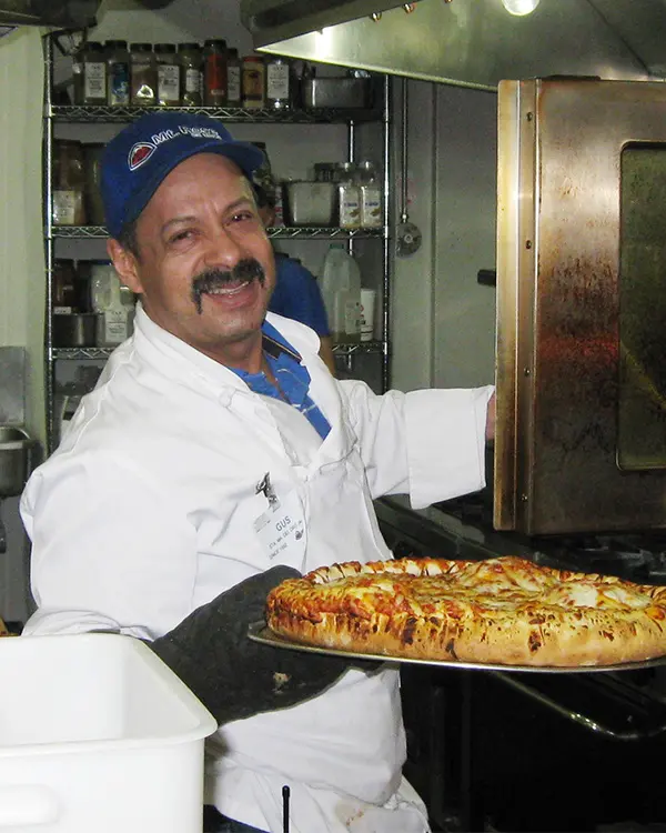 A smiling man in a white apron and blue cap removes a freshly baked pizza from an oven in a kitchen, holding the pizza tray with an oven mitt. Shelves with spices and ingredients are visible in the background, evoking après-ski vibes reminiscent of Ski Tahoe. Mt. Rose Ski Tahoe