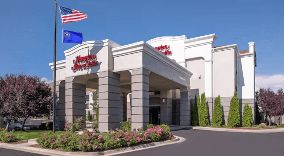 A Hampton Inn & Suites hotel building with a portico entrance, garden landscaping, and two flags—one American and one blue state flag—flying on poles against a blue sky, ideal for family ski trips or comfortable ski trip lodging. Mt. Rose Ski Tahoe