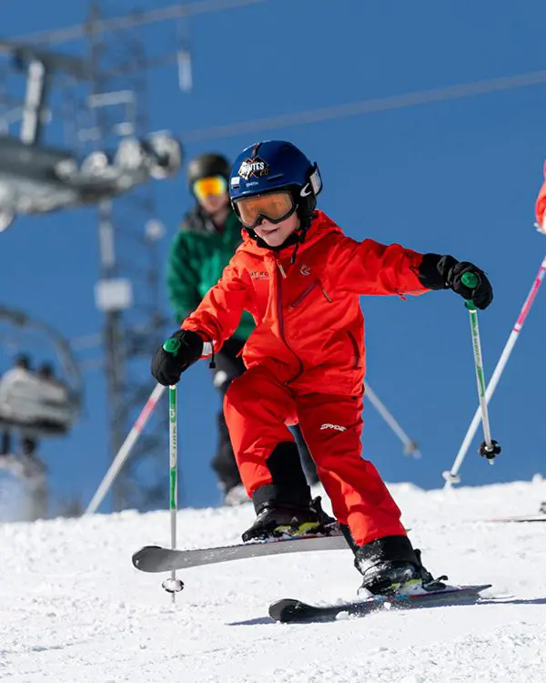 A child in a bright red ski suit and helmet skis downhill on a snowy slope at Ski Tahoe, holding ski poles. An adult and another skier are visible in the background under a clear blue sky. Mt. Rose Ski Tahoe