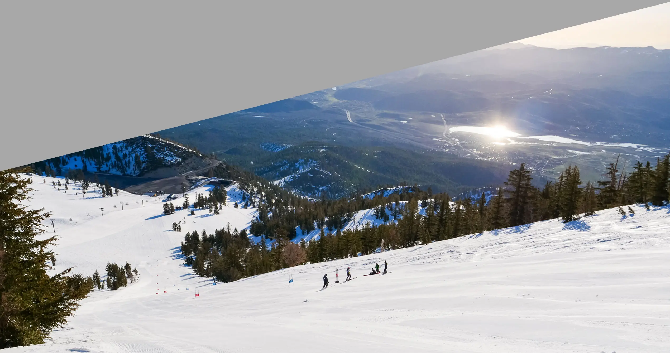 A group of skiers descends a snowy mountain slope lined with pine trees, overlooking a valley with a shimmering lake—capturing the thrill of Nevada skiing. The upper part of the image is obscured by a large gray triangle. Mt. Rose Ski Tahoe