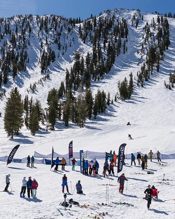 A group of people gather at the base of a snowy mountain slope with pine trees, some holding skis and snowboards. Colorful banners are displayed, and one person is skiing down the hill under a clear blue sky—true Ski Tahoe spirit. Mt. Rose Ski Tahoe