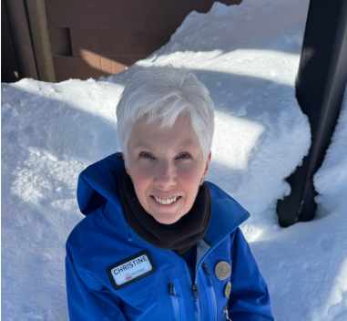 A smiling woman with short white hair, wearing a blue jacket and a name tag that reads “Christine,” stands outside in deep snow under bright sunlight, ready to welcome guests to the film festival. Mt. Rose Ski Tahoe