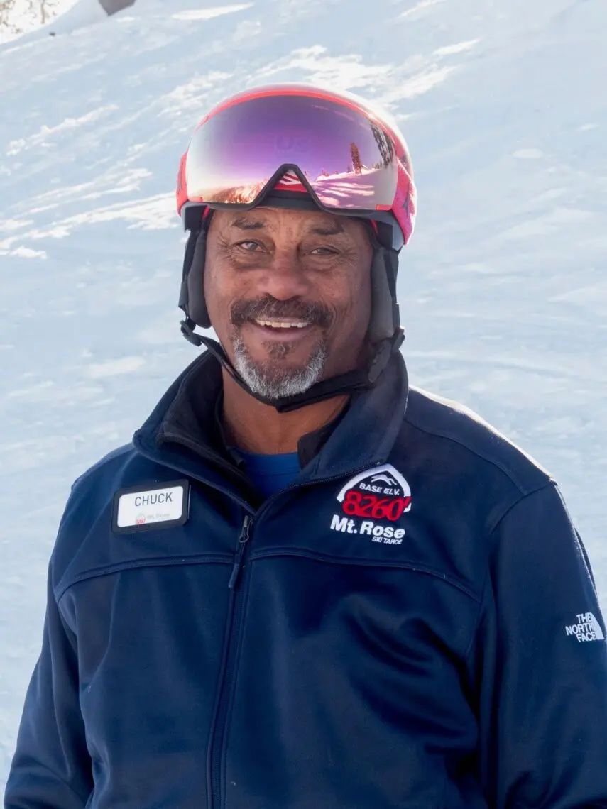 A man wearing a red ski helmet, reflective goggles, and a navy jacket with "Mt. Rose" and "Chuck" labels smiles like he's just won at a film festival, standing on a snowy slope under bright daylight. Mt. Rose Ski Tahoe