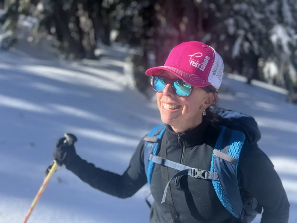A woman wearing a pink hat, blue sunglasses, and a backpack smiles while standing in a snowy forest, holding a ski pole in the sunlight, as if ready to star in her own film festival adventure. Mt. Rose Ski Tahoe