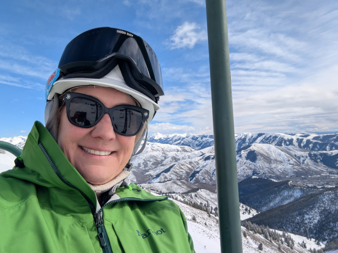 A person in a green jacket, helmet, and ski goggles smiles while riding a ski lift with snowy mountains and a partly cloudy sky in the background, as if they're ready for an adventure right out of a film festival scene. Mt. Rose Ski Tahoe