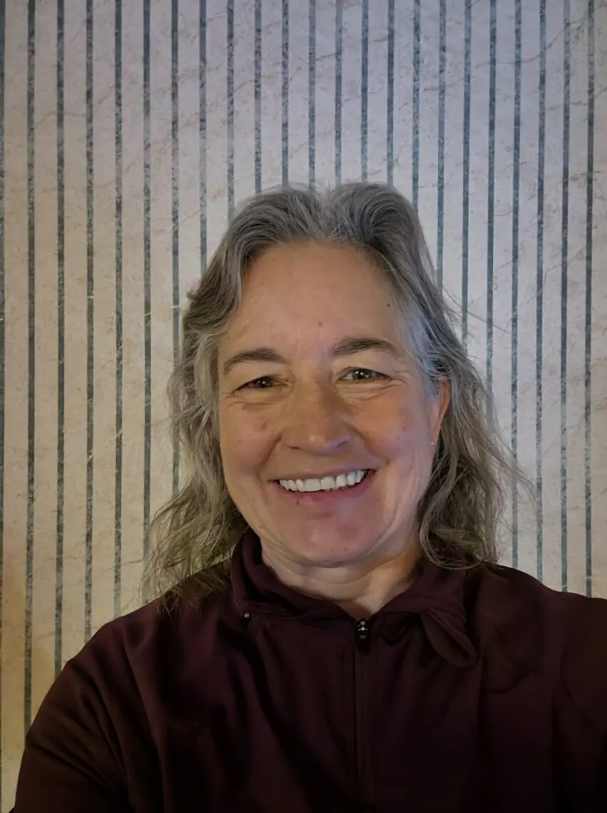 A woman with gray hair smiles at the camera, wearing a maroon jacket. She is standing in front of a wall with vertical striped lines, as if posing on the sidelines of a film festival. Mt. Rose Ski Tahoe