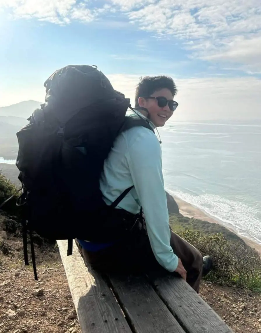 A person wearing sunglasses and a large hiking backpack sits on a bench overlooking a scenic coastal view, as if pausing before heading to a film festival, with the ocean and cliffs visible under a partly cloudy sky. Mt. Rose Ski Tahoe