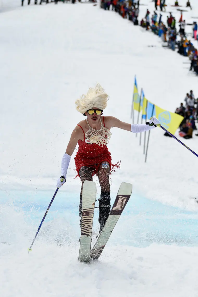 A skier wearing a blonde wig, red dress, and white gloves skis downhill on snow, with a crowd of spectators and flags in the background. The skier appears to be participating in a fun or costume event. Mt. Rose Ski Tahoe