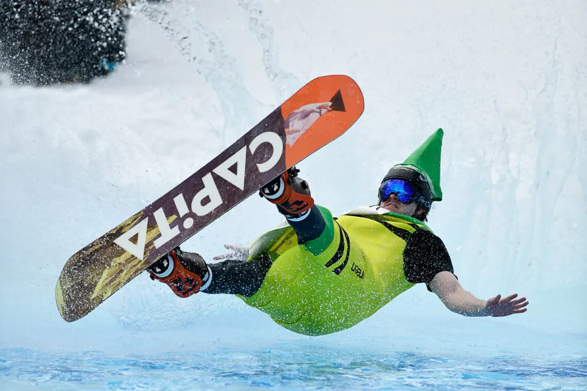 A snowboarder wearing a bright costume and green cone hat falls backward into a pool of water, creating a splash, during a snowboarding event. Mt. Rose Ski Tahoe
