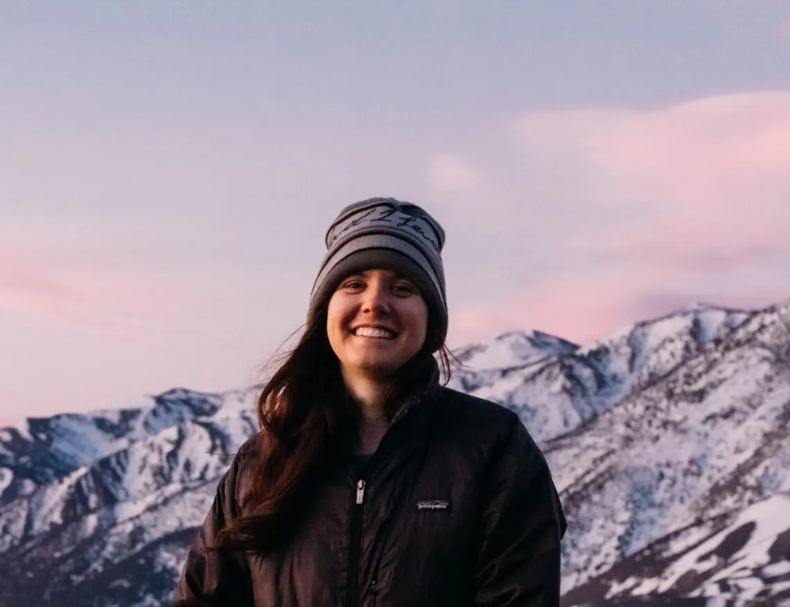 A woman wearing a beanie and jacket smiles while standing outdoors, as snow-covered mountains and a pinkish sky create a scene reminiscent of a film festival backdrop. Mt. Rose Ski Tahoe