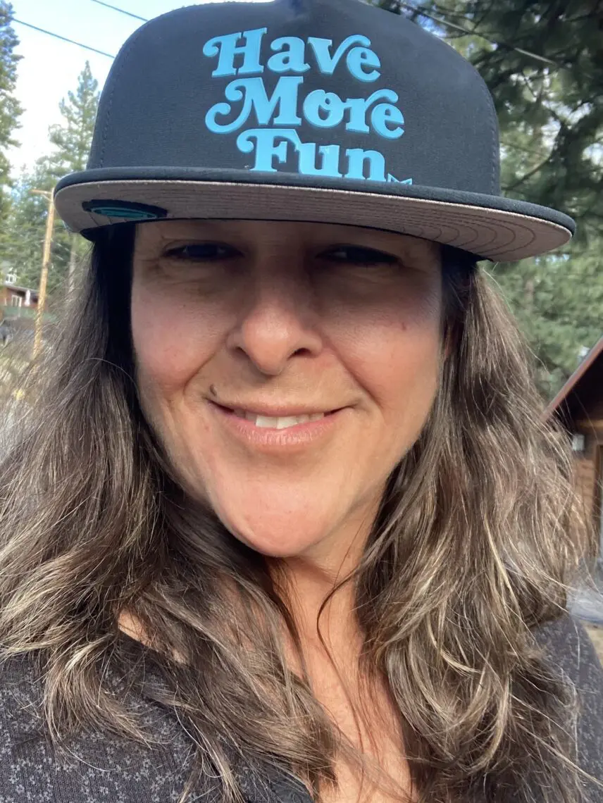 A woman with long brown hair smiles outdoors at a film festival, wearing a black cap with blue text that reads "Have More Fun." Trees and wooden buildings are visible in the background. Mt. Rose Ski Tahoe