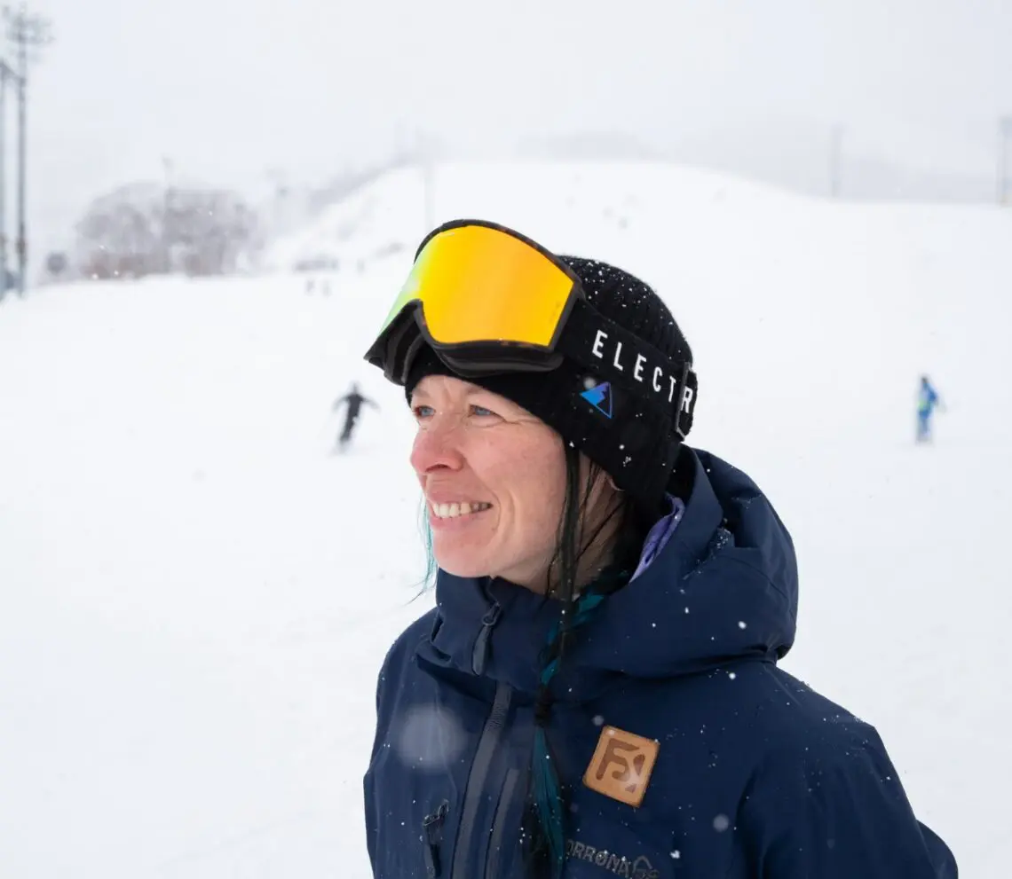 A woman wearing a black ski hat, yellow ski goggles, and a blue jacket smiles while standing on a snowy slope, as if ready for a film festival set in the mountains, with skiers in the background on a cloudy, snowy day. Mt. Rose Ski Tahoe