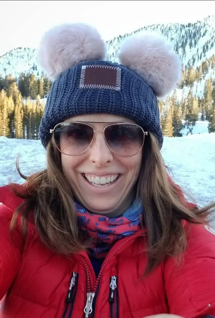 A woman wearing sunglasses, a red winter jacket, and a blue knit hat with two fluffy pom-poms smiles outdoors in a snowy mountain landscape, capturing the spirit of a film festival set against breathtaking alpine scenery. Mt. Rose Ski Tahoe