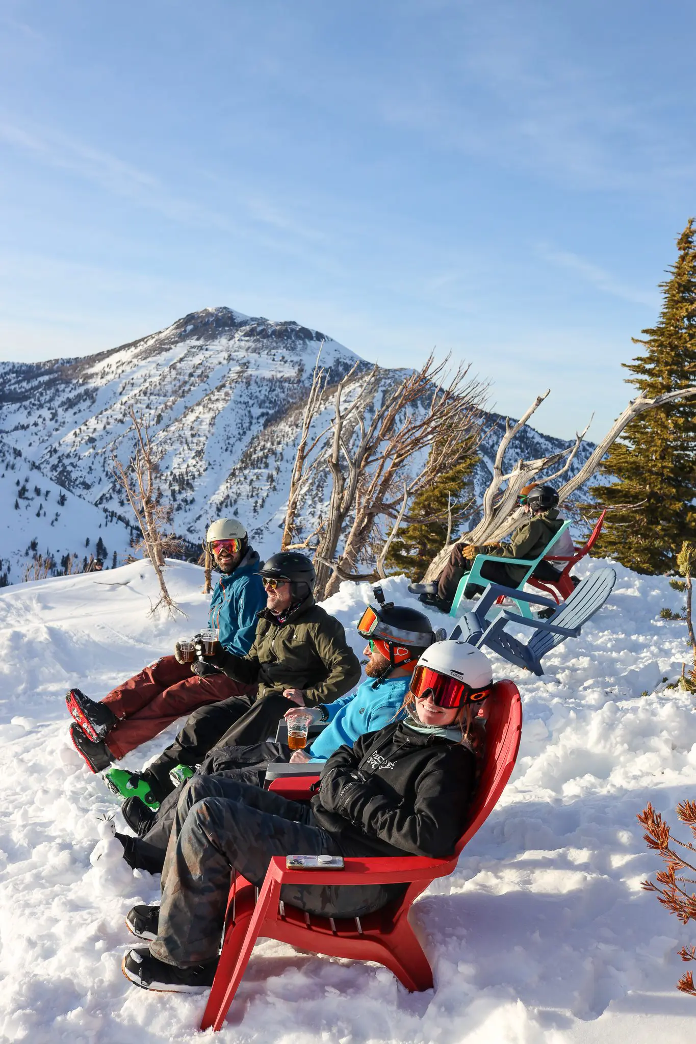Five people in ski gear sit in colorful chairs on a snowy mountain slope, relaxing with drinks. Snow-covered peaks and trees are visible in the background under a clear blue sky. Mt. Rose Ski Tahoe
