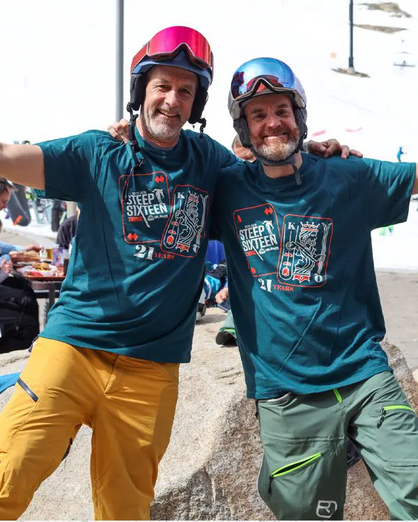 Two smiling men in ski helmets and matching “Steep Sixteen” T-shirts pose together at a snowy Ski Tahoe resort, each with an arm around the other. One wears yellow pants, the other green. Mt. Rose Ski Tahoe