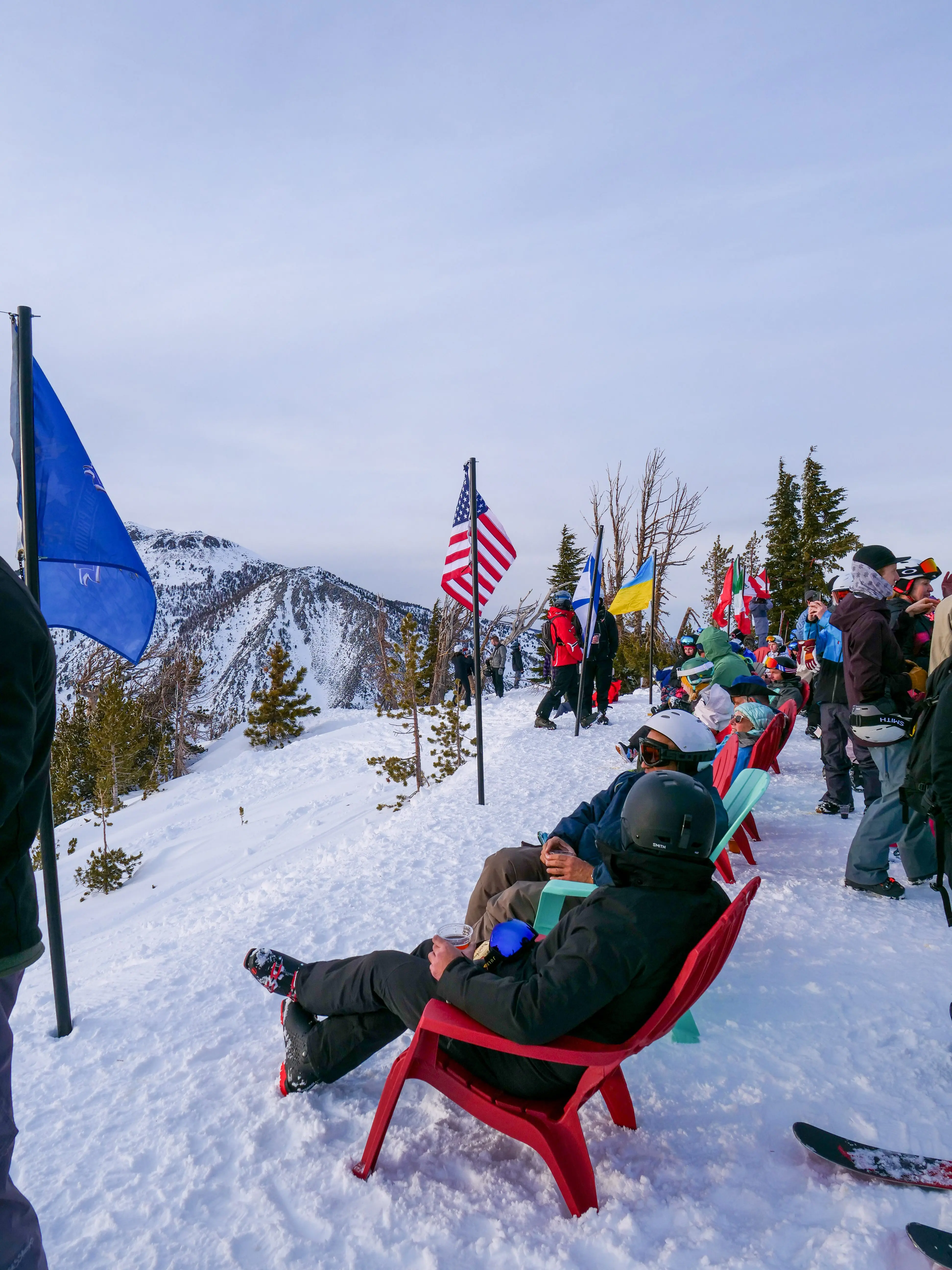 People in ski gear sit on colorful chairs on a snowy mountain slope, with flags including the American flag nearby. Snow-covered mountains and pine trees are visible in the background under a cloudy sky. Mt. Rose Ski Tahoe